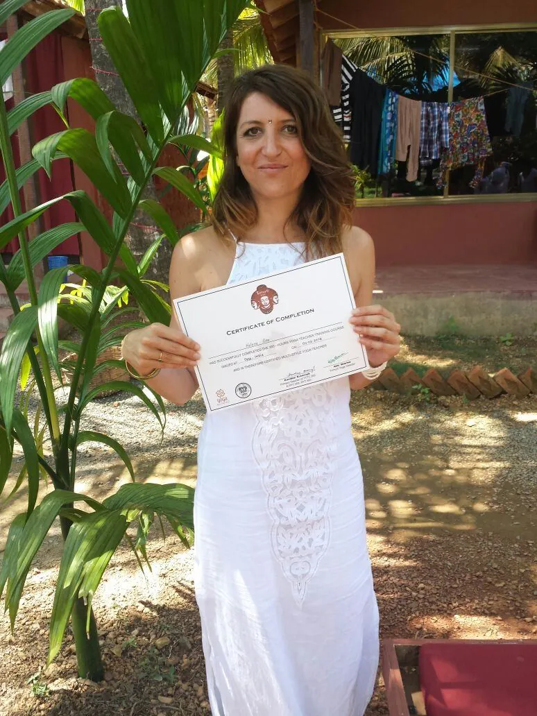 Helena Ona en robe blanche, tenant son certificat de professeure de yoga, dans le jardin du centre Trimurti à Goa
