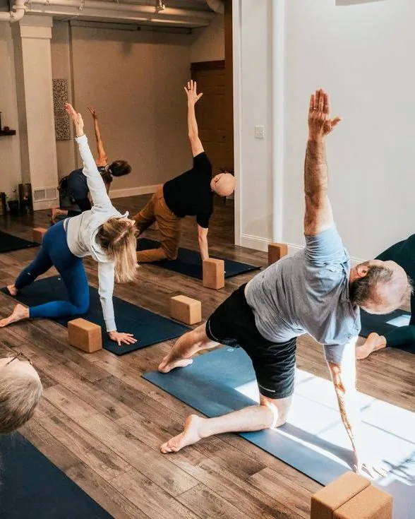 Un groupe d'adultes en équilibre sur un genou, un bras tendu vers le plafond, pendant un cours de yoga en salle