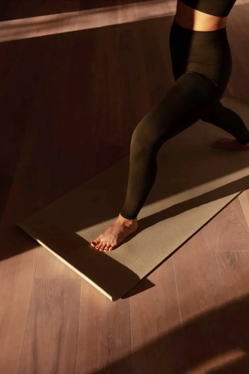 Une jambe en fente, le pied posé sur un tapis de yoga, dans la lumière rasante d'une salle au parquet